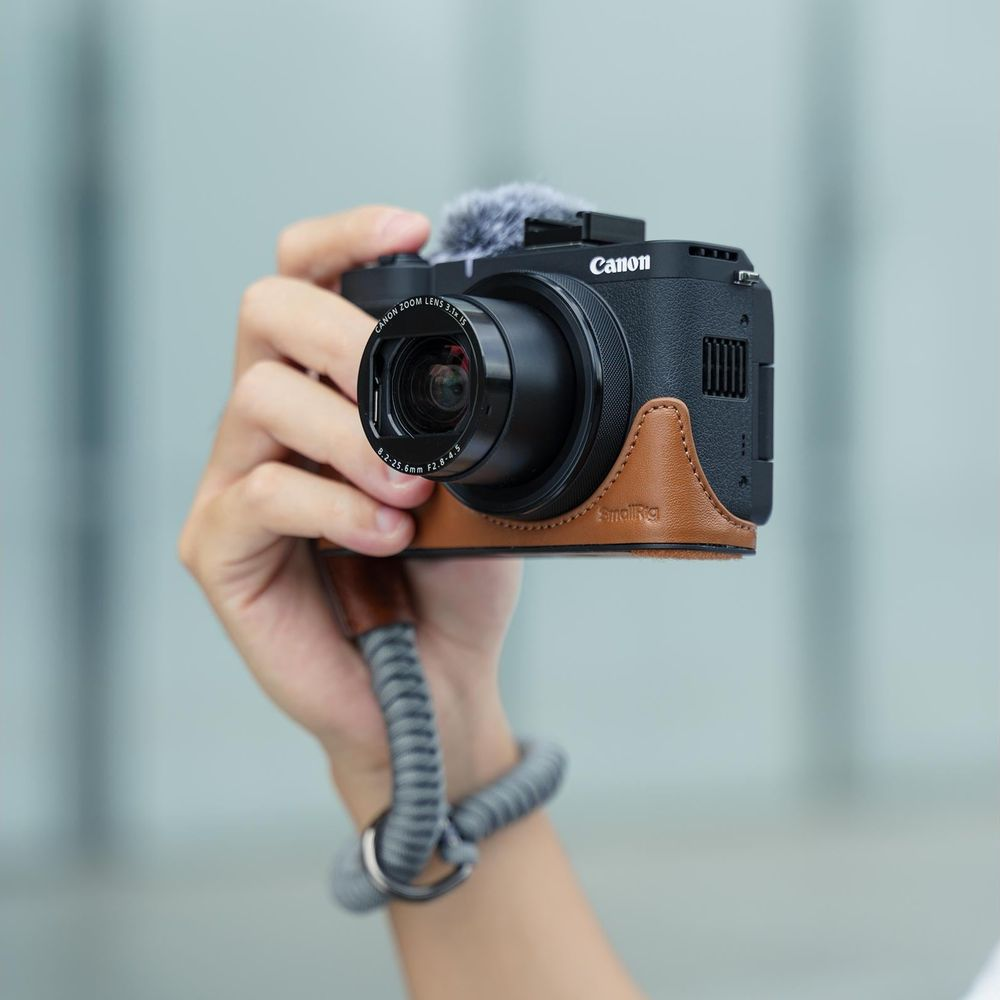 A hand holds a black Canon camera with brown leather case and microphone.