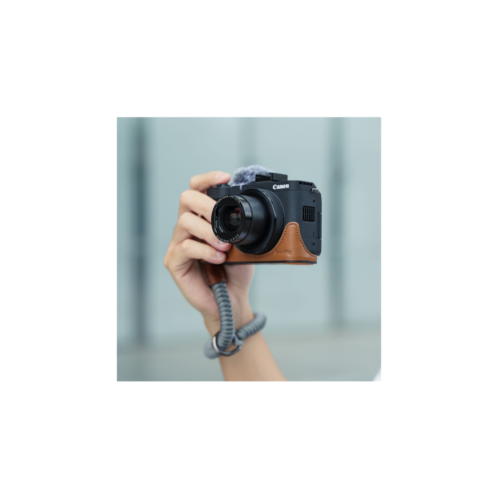 A hand holds a black Canon camera with a brown leather case and wrist strap.