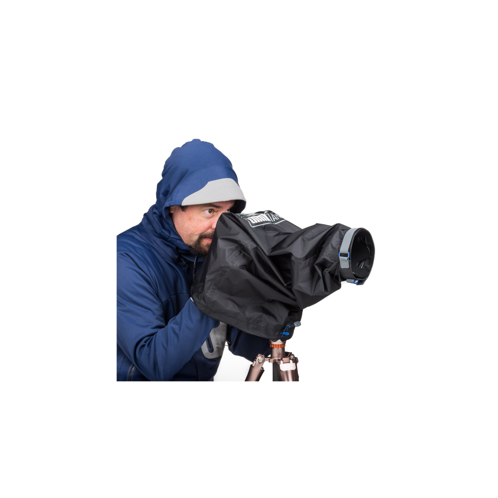 Man in blue hooded jacket peers through camera with rain cover on tripod.