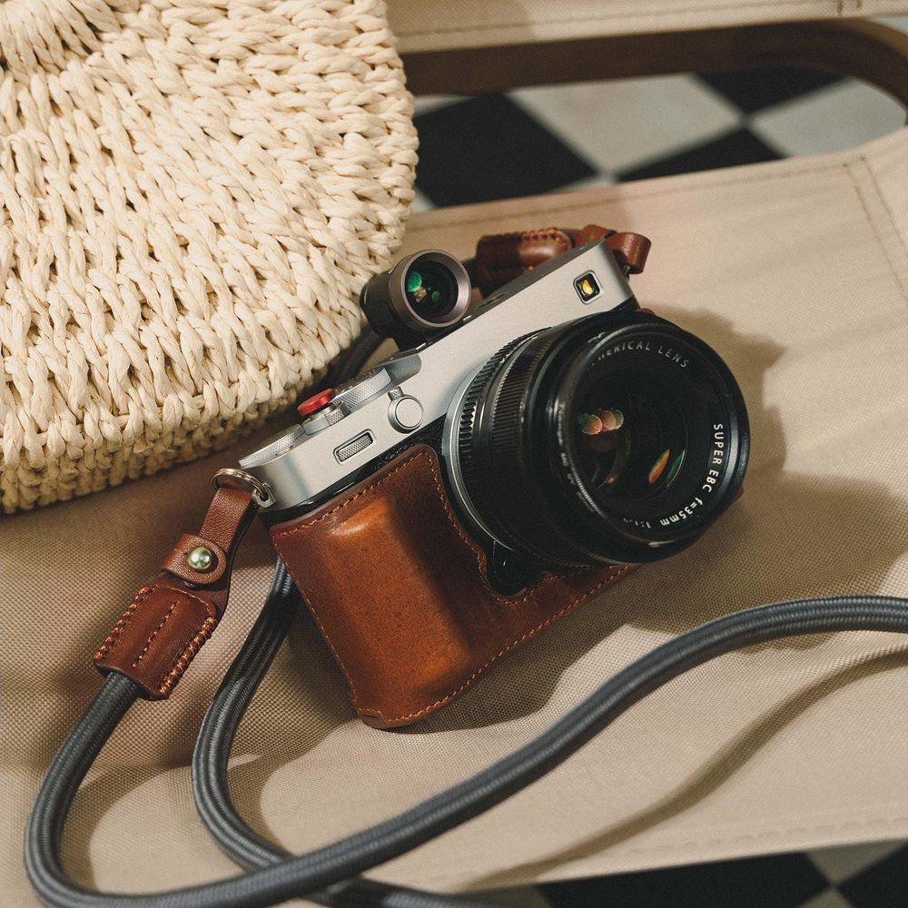 Zilveren camera met bruine lederen hoes en riem, naast een strooien tas.
