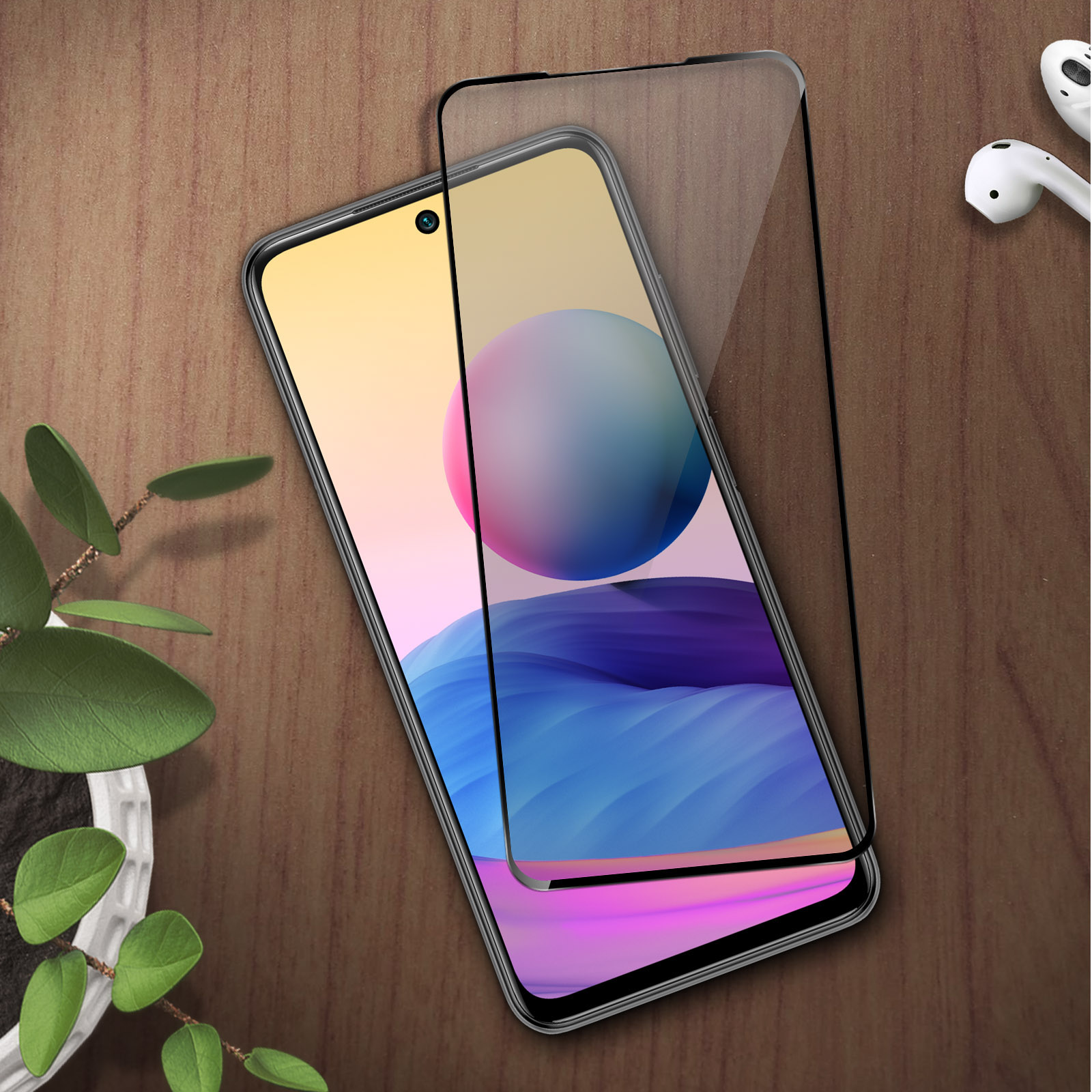 Uno smartphone con proteggi schermo su tavolo in legno, accanto a auricolari e pianta.