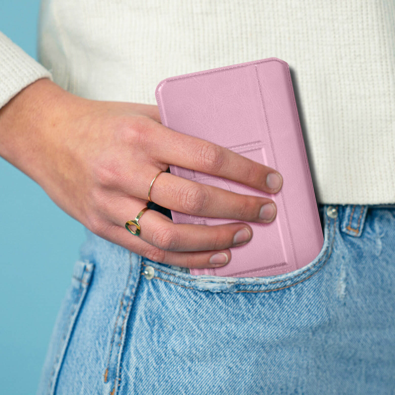 Une main en jean place un étui rose pour tablette dans une poche. Des bagues dorées ornent les doigts.