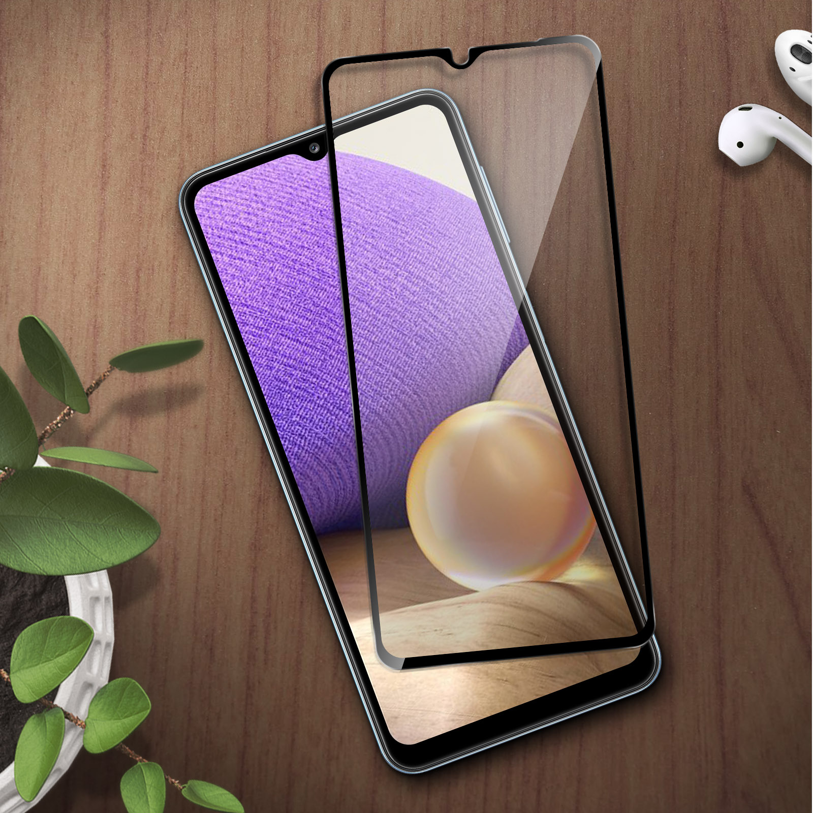 Een smartphone met screenprotector op een houten tafel, naast een plant en oordopjes.