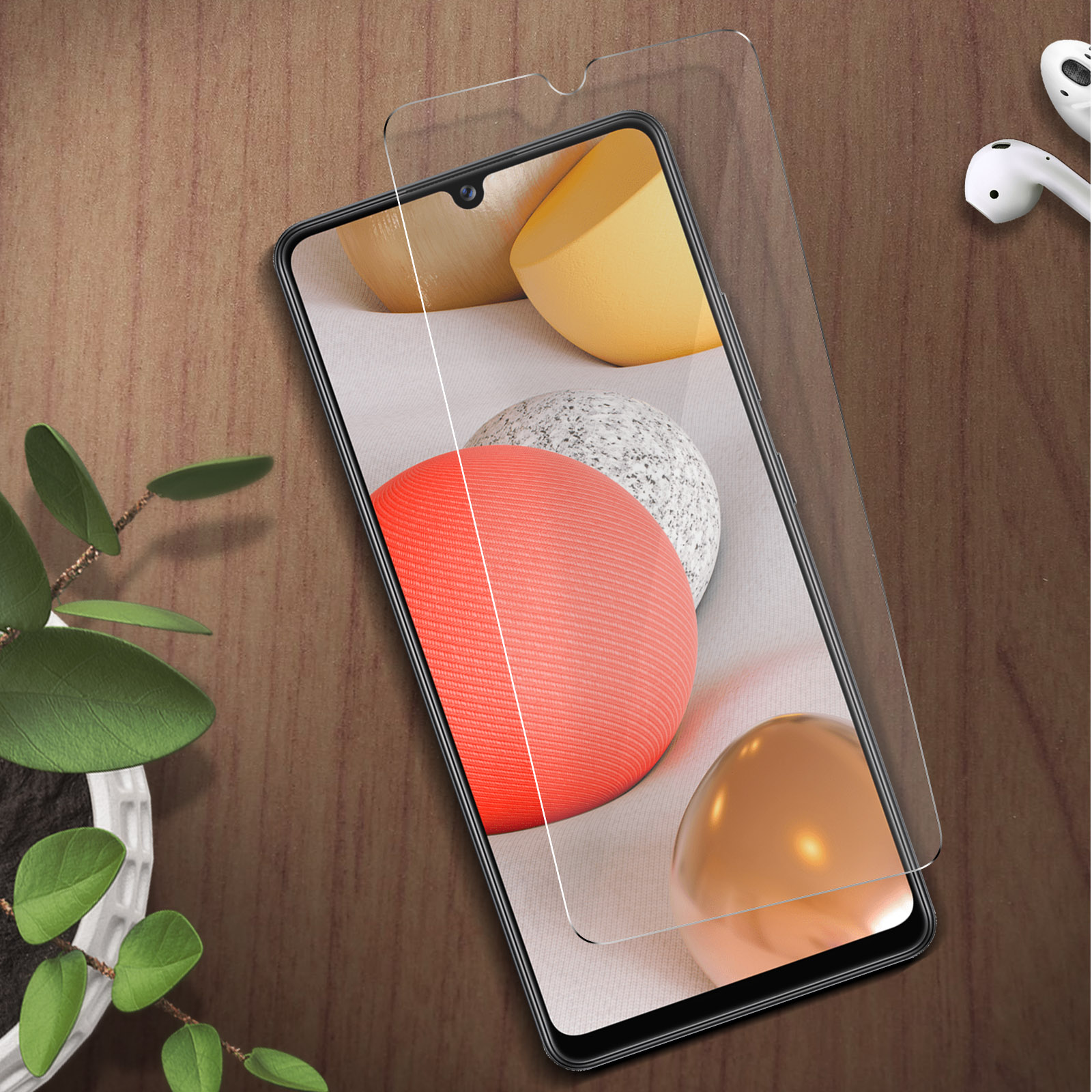 Uno smartphone con pellicola protettiva, auricolari bianchi e una pianta vicina.