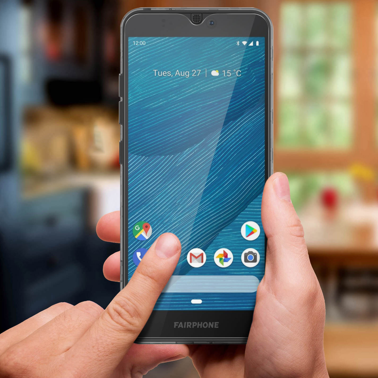 Une main tient un Fairphone avec un fond bleu et des icônes d'applications. 12h00, Mar 27.