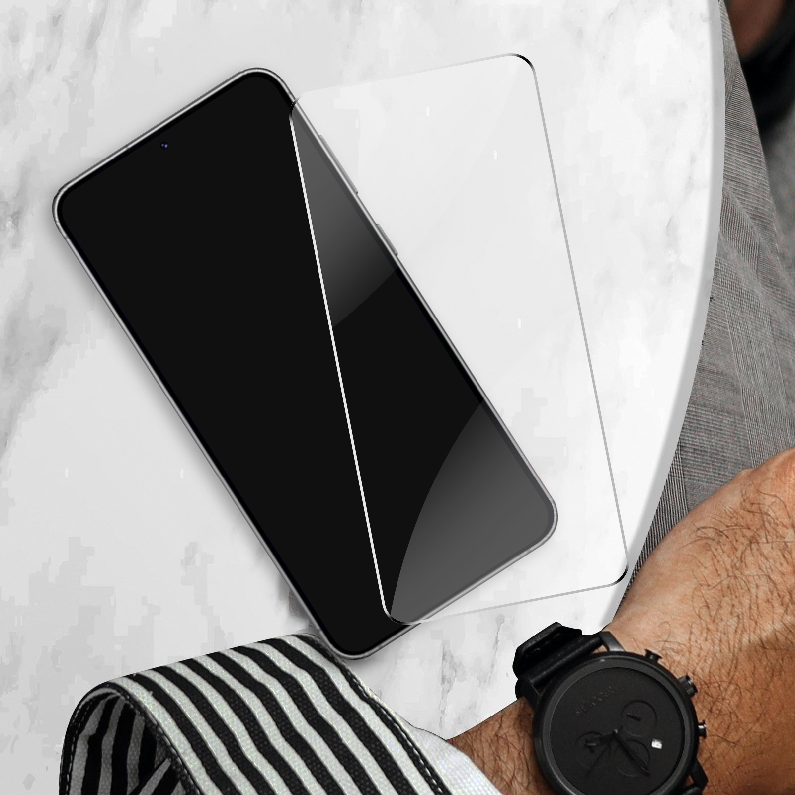 Un smartphone noir avec un protecteur d'écran sur une surface en marbre. Une montre est visible.
