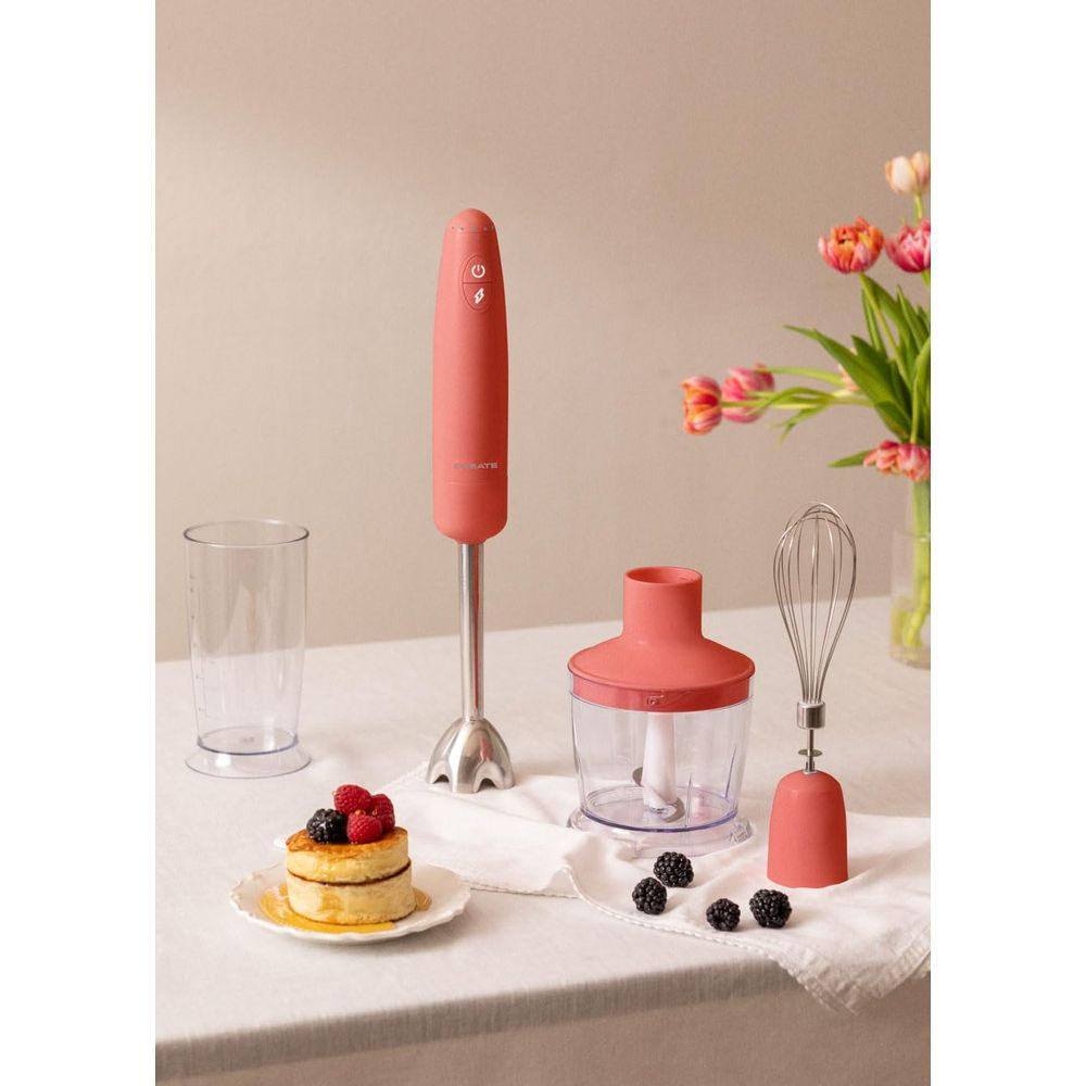 Una batidora de mano coral con accesorios está sobre una mesa junto a un vaso y un postre.