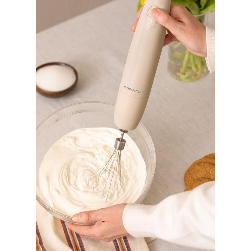 Una mano usa una batidora de mano beige para batir crema en un bol de vidrio. Hay azúcar cerca.
