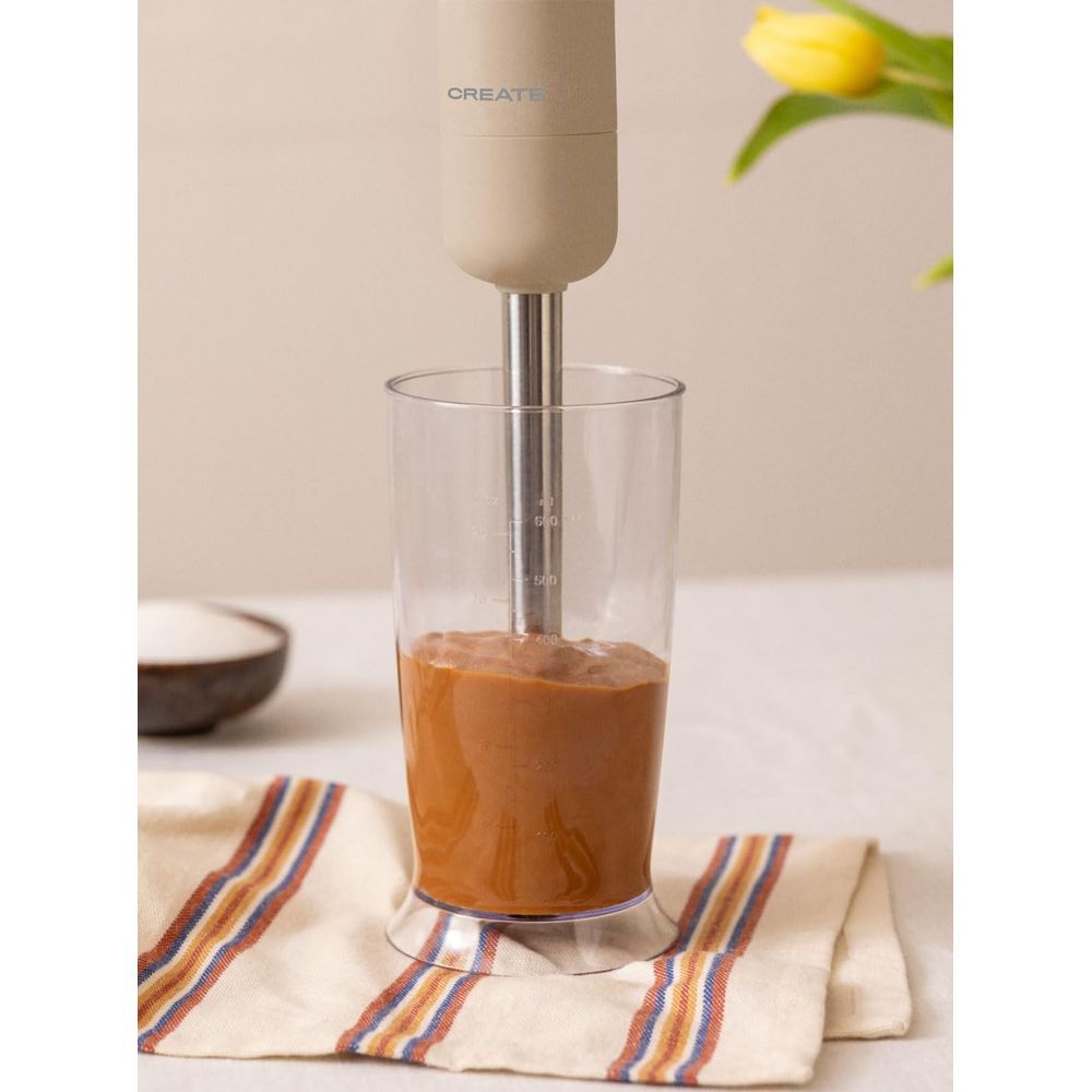 Una batidora de inmersión mezcla un líquido marrón en un vaso medidor. Hay una tela al lado.