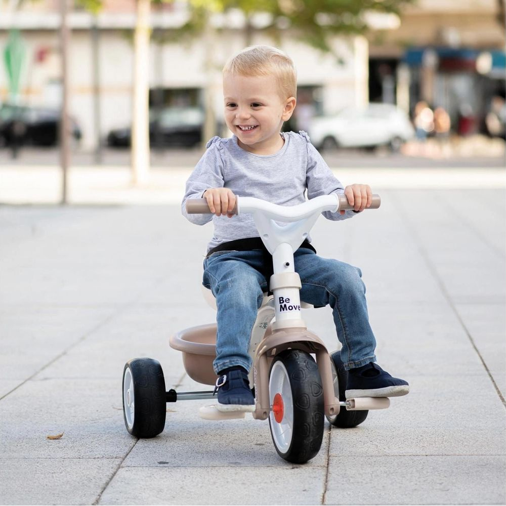 Un bambino sorridente guida un triciclo beige e bianco su un marciapiede pavimentato all'aperto.