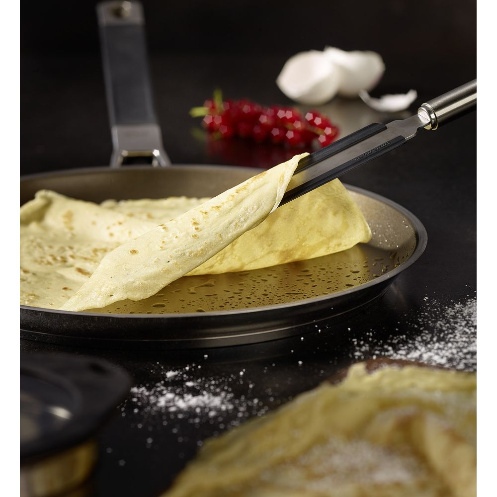 Ein Pfannkuchen wird mit einer Zange aus einer Pfanne genommen. Rote Johannisbeeren und eine weiße Zwiebel liegen daneben.