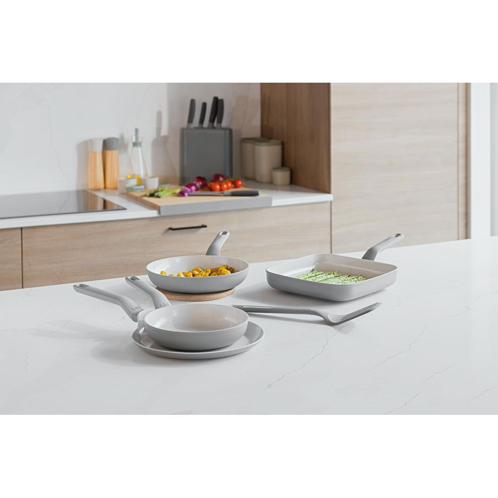 Grey cookware set on a white countertop with kitchen background. Includes pans with food.