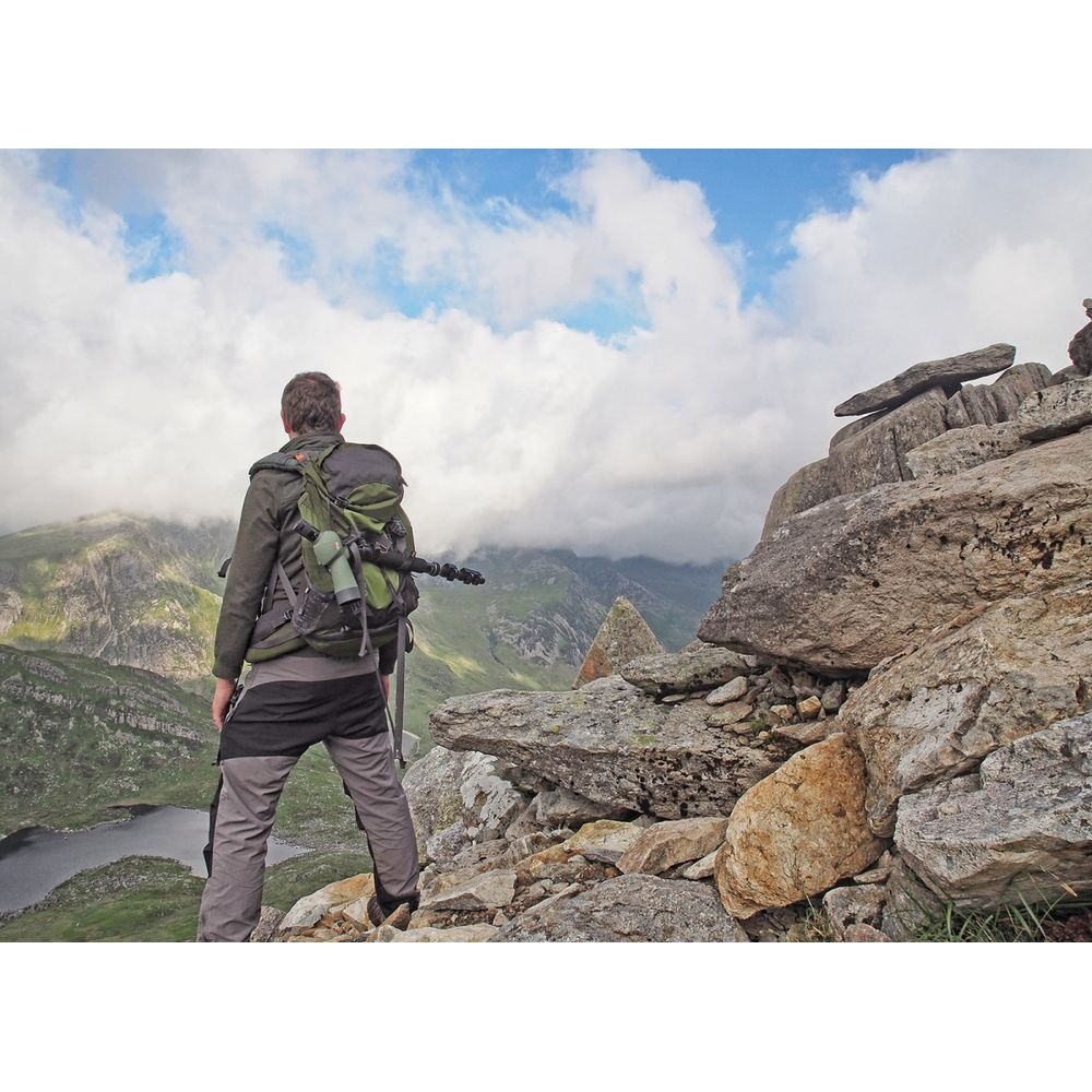 Un escursionista con zaino si erge su una cima rocciosa affacciata su montagne e nuvole.