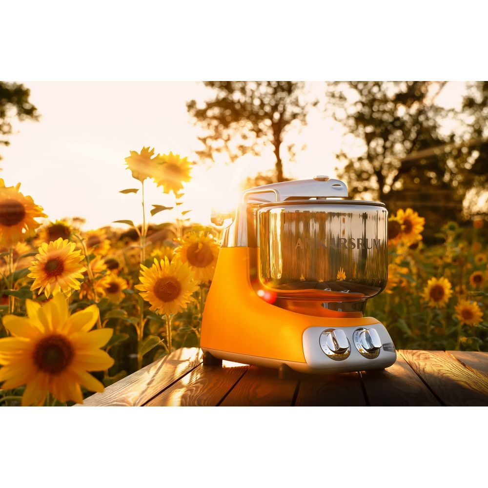 Ein orangefarbener Küchenmixer steht auf einem Holztisch vor einem Sonnenblumenfeld bei Sonnenuntergang.
