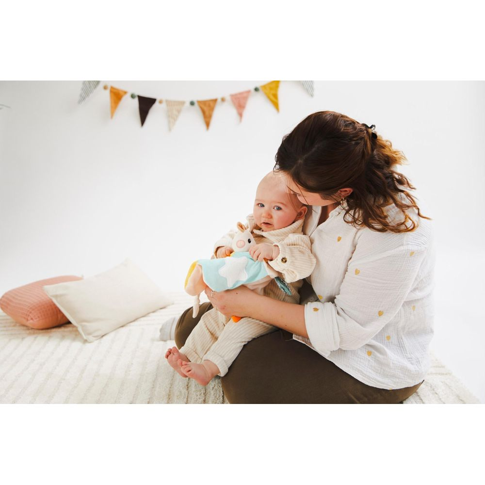 Une femme tient un bébé tenant un doudou. Des fanions colorés pendent en arrière-plan.