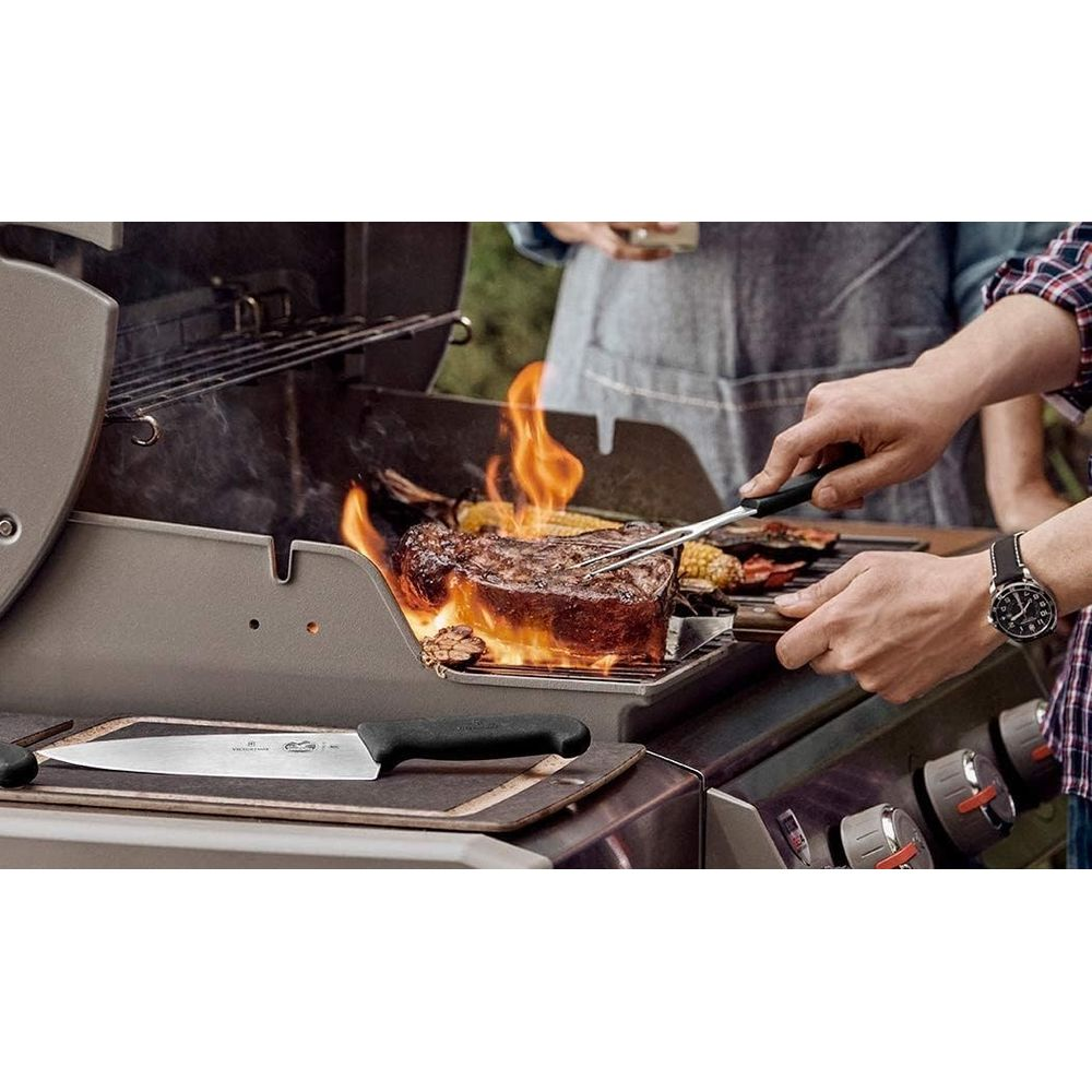 Ein dicker Steak wird über Flammen gegrillt, mit Mais und Gemüse in der Nähe. Ein Koch wendet das Steak mit einer Zange.