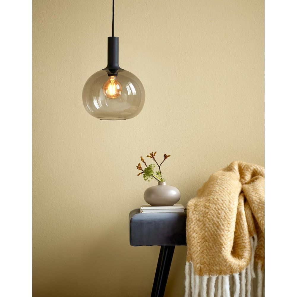Une lampe suspendue ronde en verre fumé surplombe une petite table avec vase et jeté.