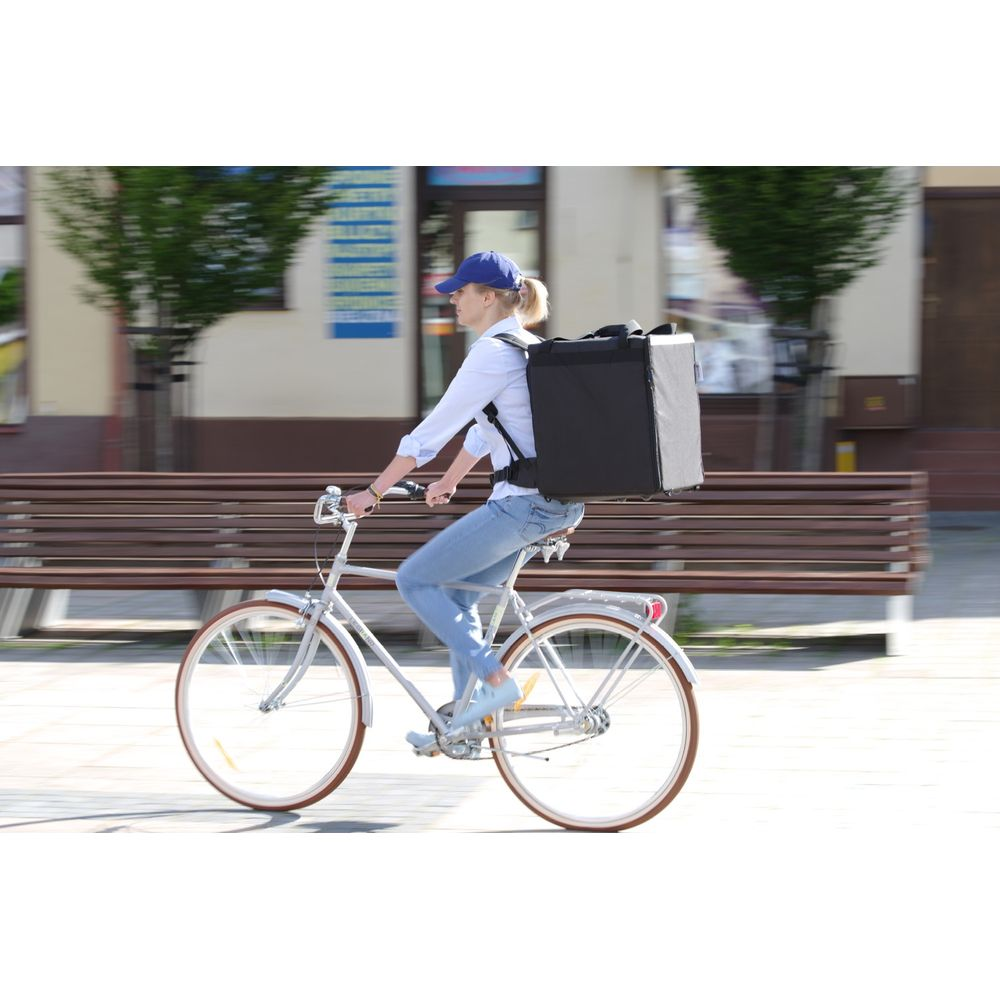 Donna con grande borsa nera da consegna sulla schiena pedala bici bianca in strada.