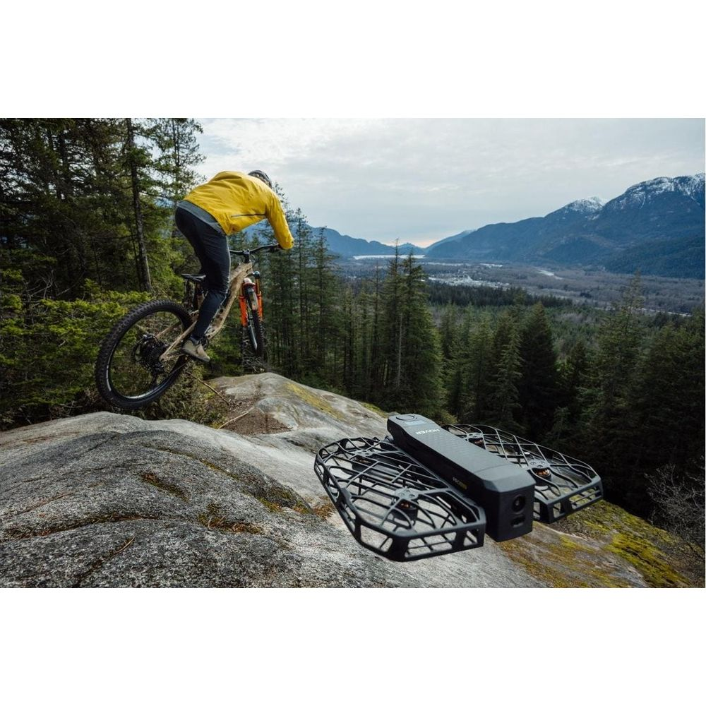 Ein Mountainbiker in einer gelben Jacke springt über Felsen, eine Drohne im Vordergrund.