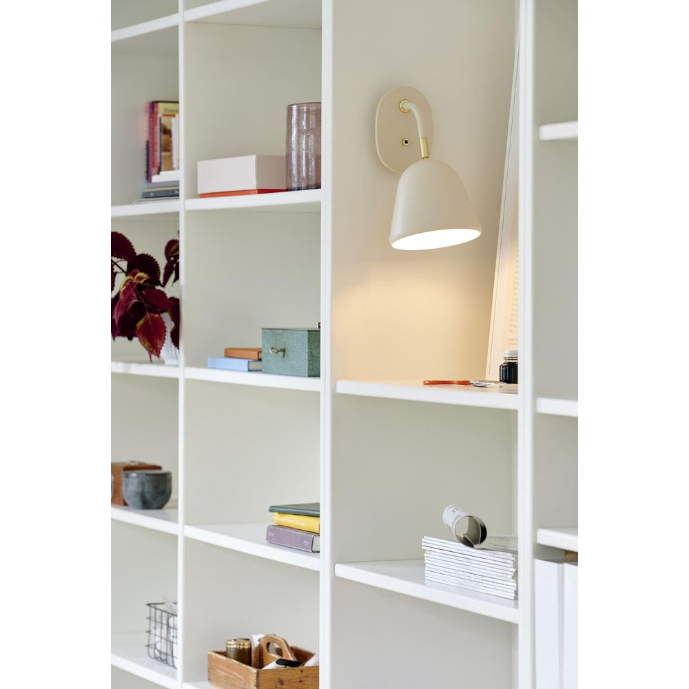Eine weiße Wandleuchte beleuchtet ein Regal mit Büchern, einer Pflanze und Deko-Boxen.