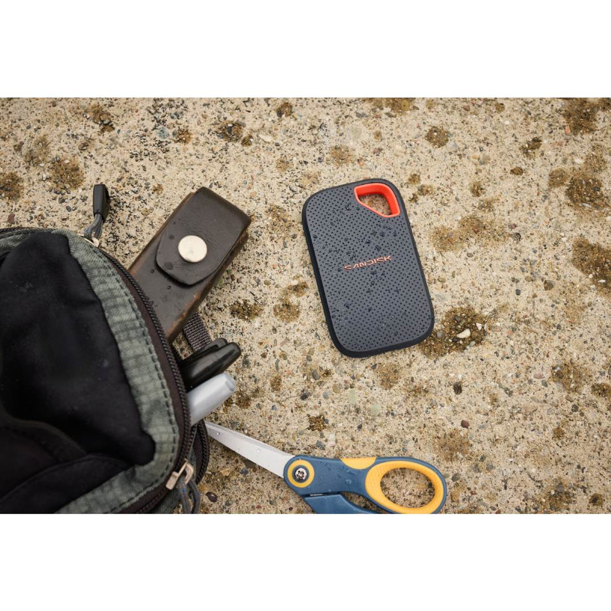 Turuncu kenarlı koyu gri Sandisk sürücüsü, çanta ve makasın yanında betonda duruyor.