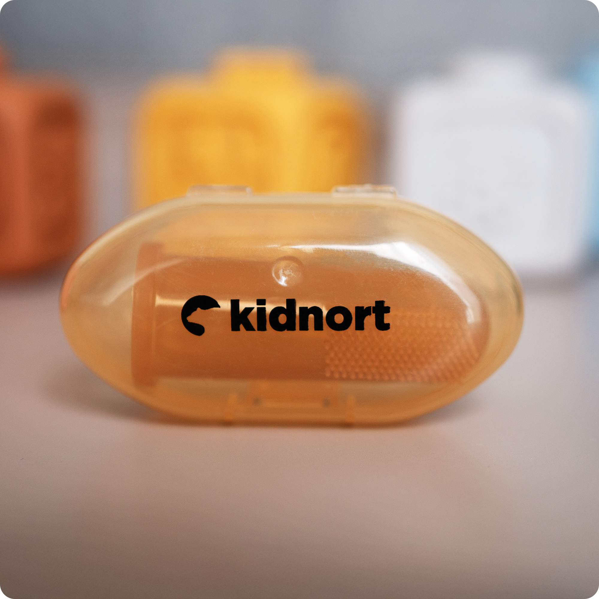 Przezroczyste pomarańczowe etui z logo 'kidnort', rozmyte kolorowe obiekty w tle.
