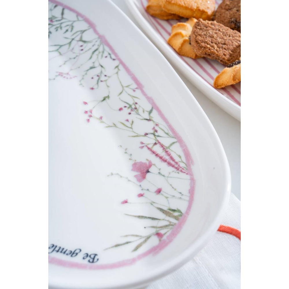 Pembe çiçekli ve 'Be gentle' yazılı beyaz oval tabak. Arkasında kurabiyeli başka bir tabak.