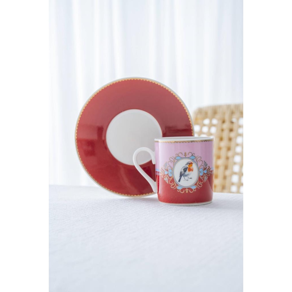 Beyaz masa örtüsü üzerinde kırmızı fincan tabağı ve kuş desenli pembe kupa.