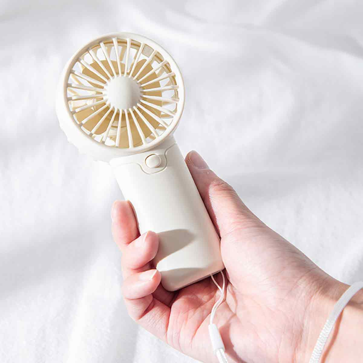 Eine Hand hält einen kleinen, cremefarbenen tragbaren Ventilator vor einem weißen, strukturierten Hintergrund.