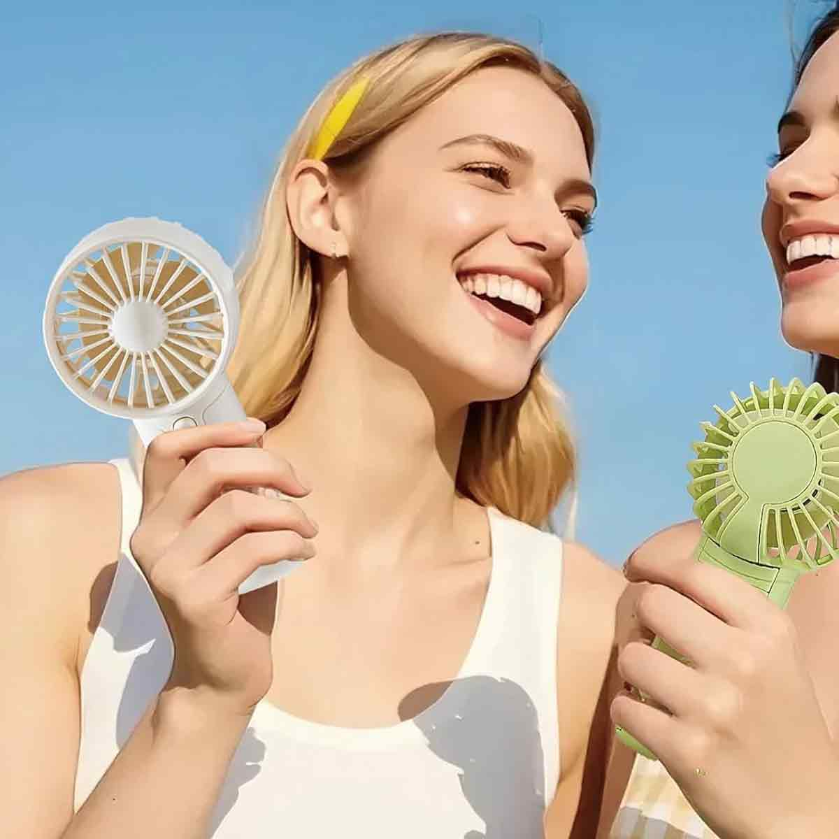 Zwei Frauen lachen und halten tragbare Ventilatoren vor blauem Himmel.