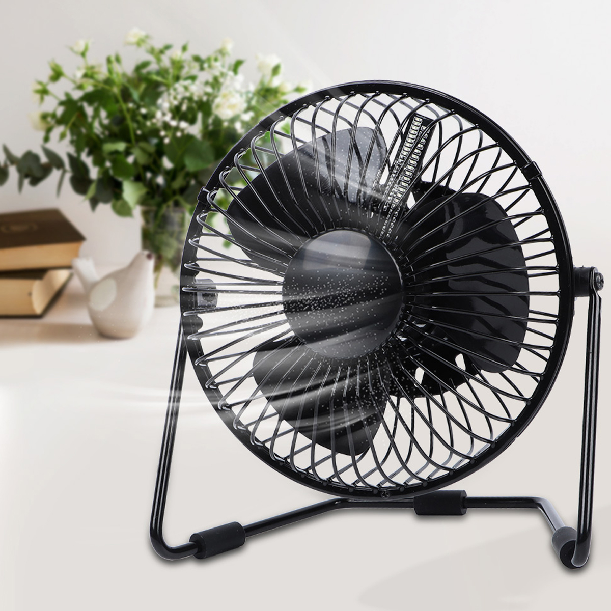 Ein schwarzer Schreibtischventilator mit rotierenden Flügeln auf einer weißen Oberfläche, mit Büchern und Pflanzen im Hintergrund.