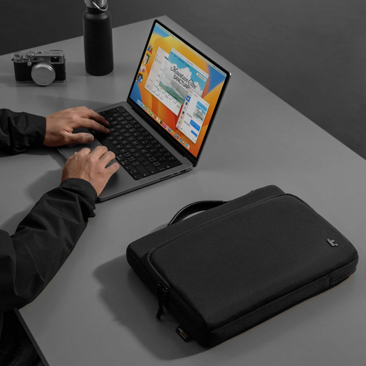 Persona digita su laptop accanto a borsa, macchina fotografica e bottiglia.