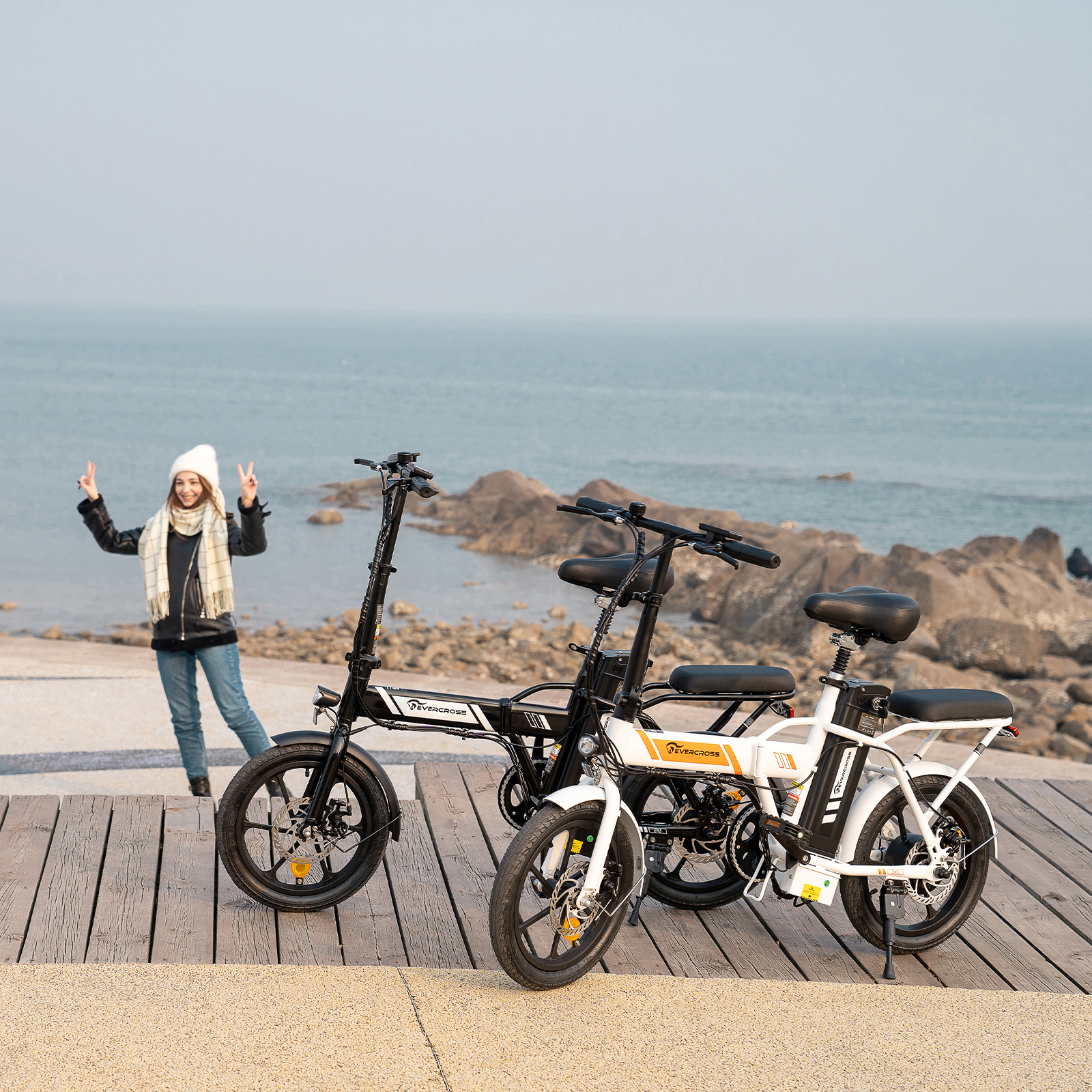 Vrouw met twee fietsen aan zee. Een vrouw maakt vredestekens naast twee e-bikes aan zee.