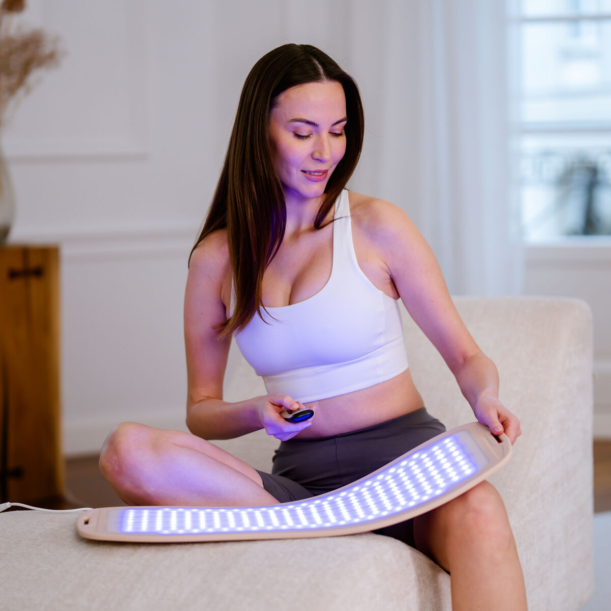 Mujer con top blanco y shorts grises sostiene control, sentada con dispositivo de fototerapia azul.