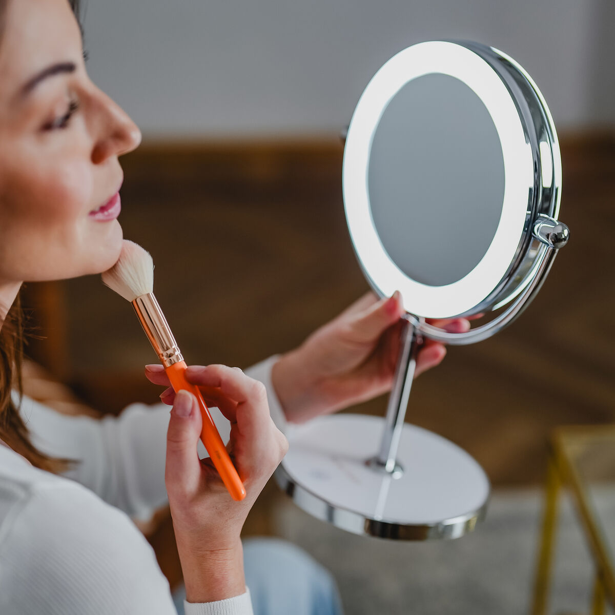 Eine Frau trägt mit einem Pinsel Make-up vor einem beleuchteten Spiegel auf.