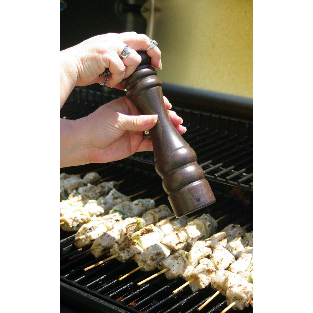 Holzmühle würzt Hähnchenspieße auf dem Grill. Hände halten die Mühle.