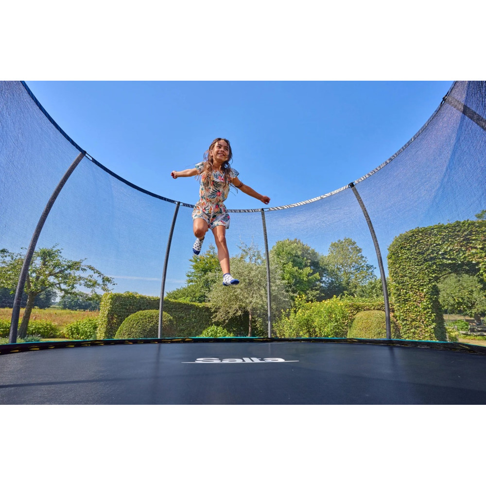 Młoda dziewczyna w kwiecistej kombinezonce skacze wysoko na trampolinie na zewnątrz pod błękitnym niebem.