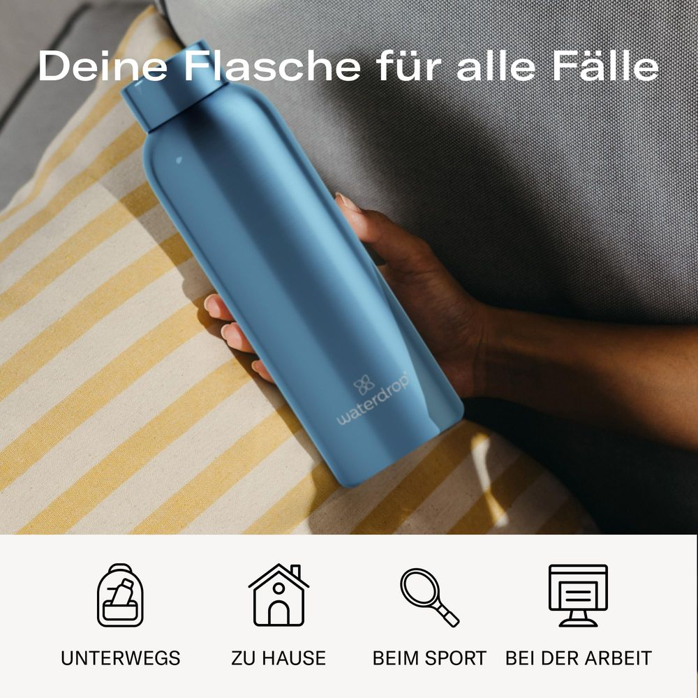 Eine blaue Waterdrop-Flasche wird von einer Hand gehalten, neben Kissen und Bettdecke.