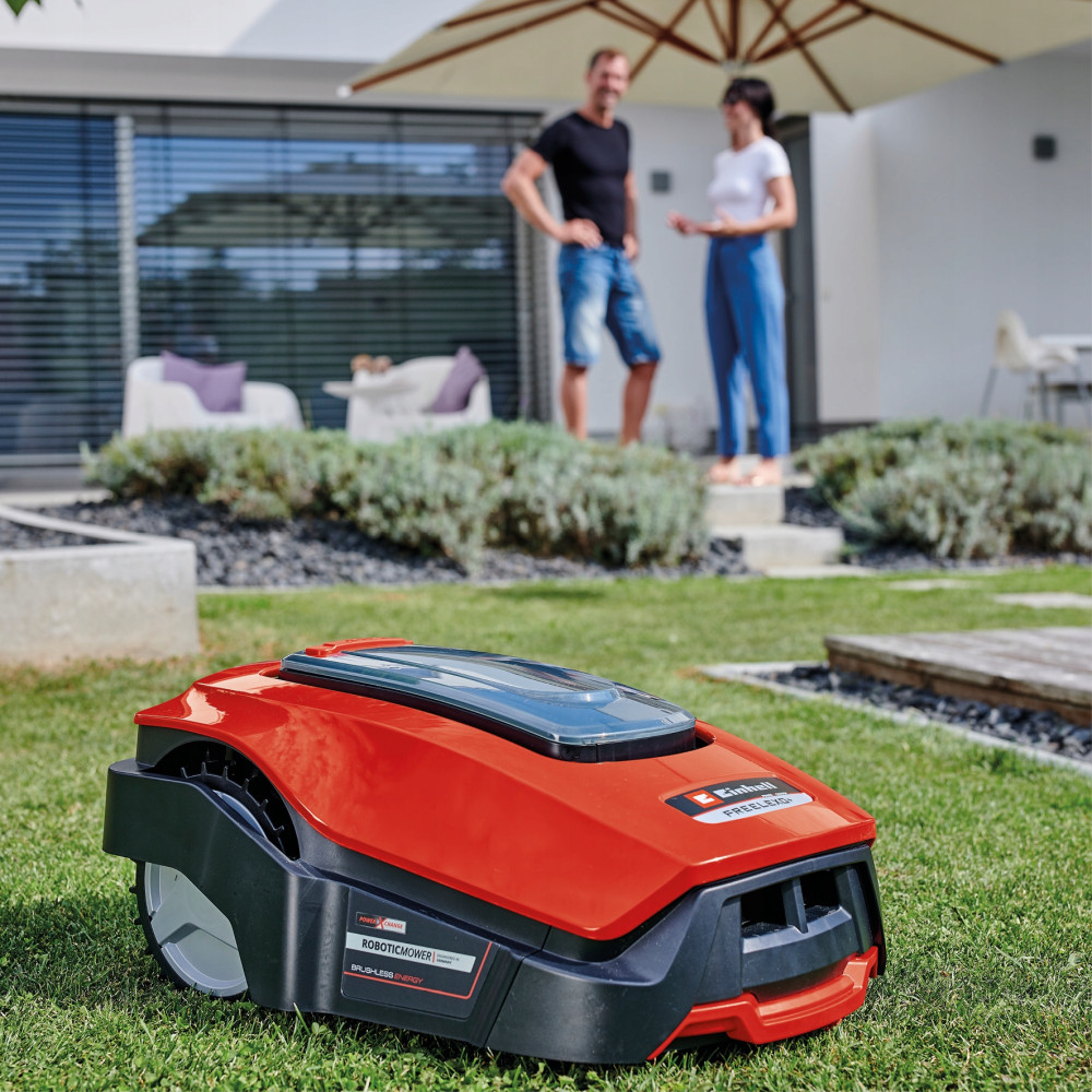 Czerwony robot koszący stoi na trawie przed domem, za nim stoi para.