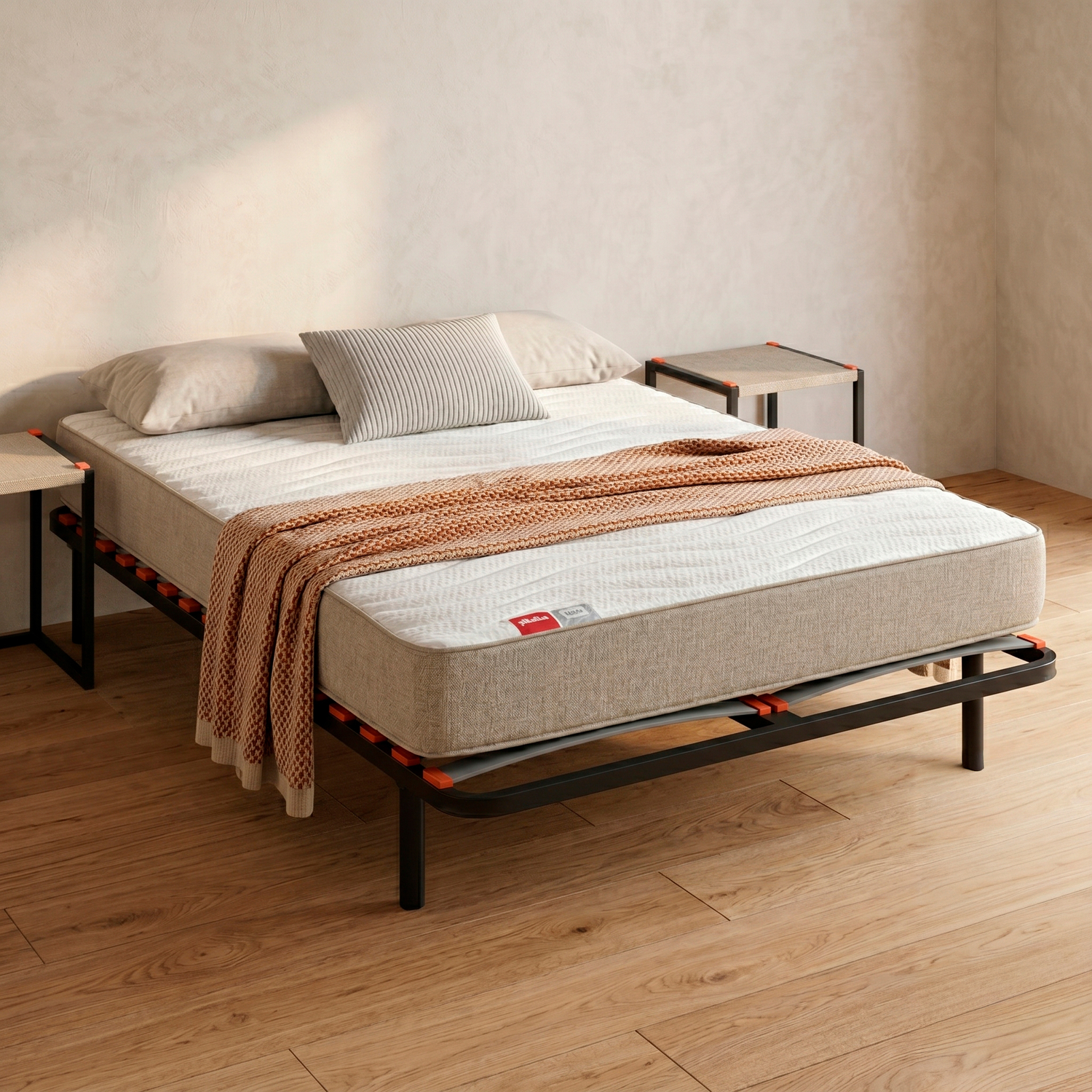 Una cama con colchón, almohadas y manta sobre un marco metálico y suelo de madera.