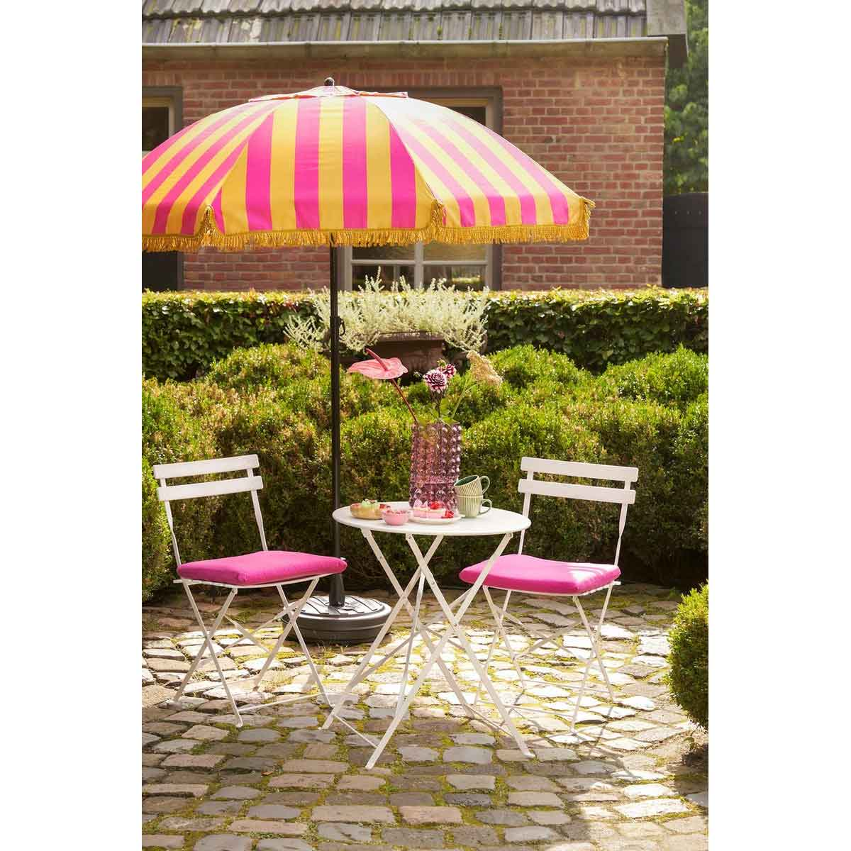 A white table and two chairs with pink cushions under a striped umbrella.