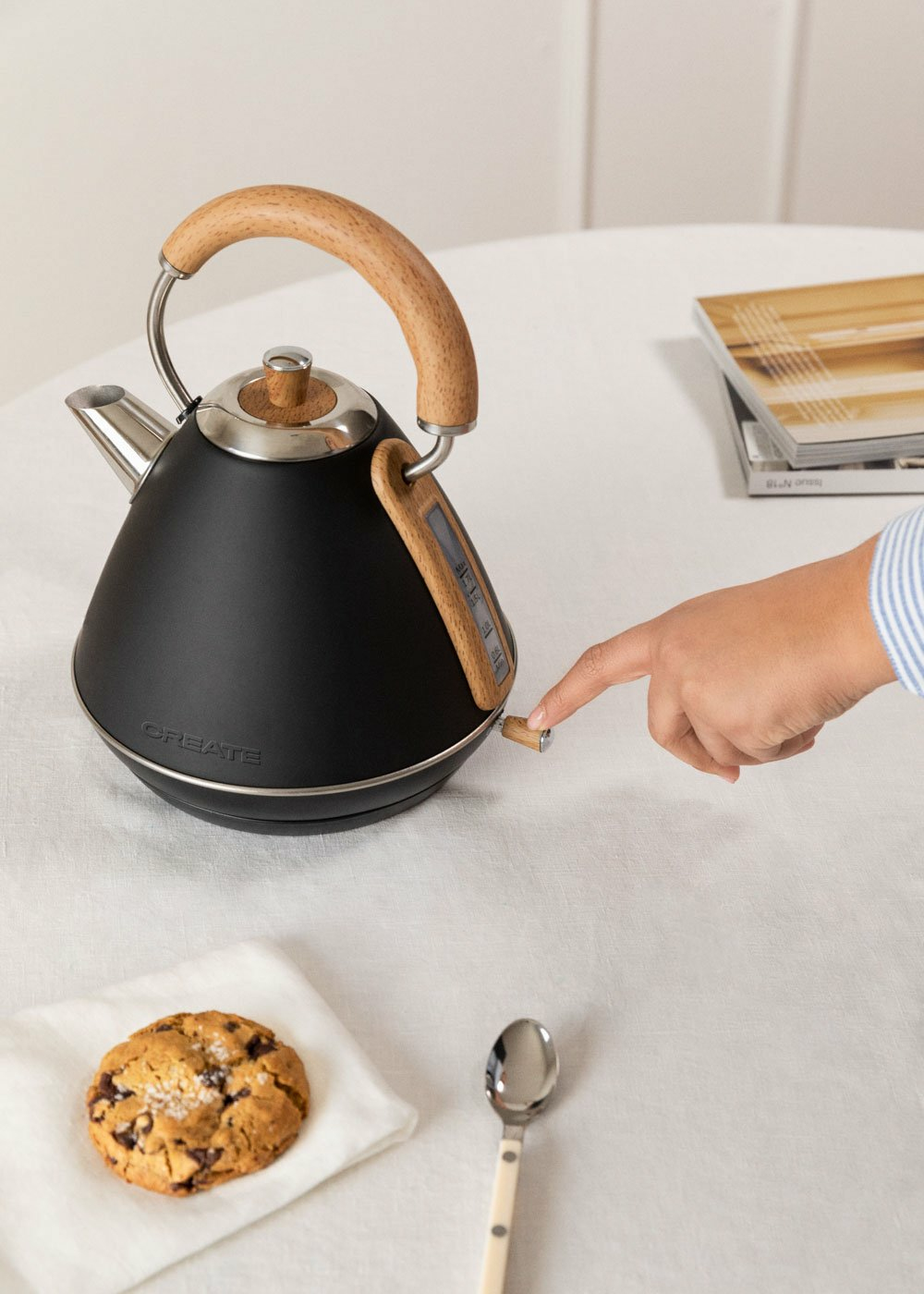 Un hervidor eléctrico negro con mango y detalles de madera sobre un mantel blanco.