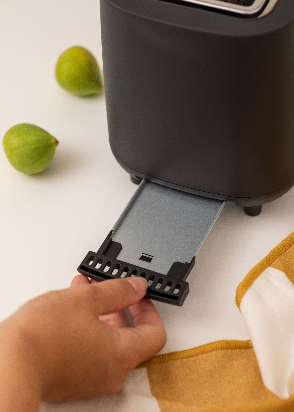 Une main retire le bac à miettes d'un grille-pain sombre. Deux figues vertes sont à proximité.