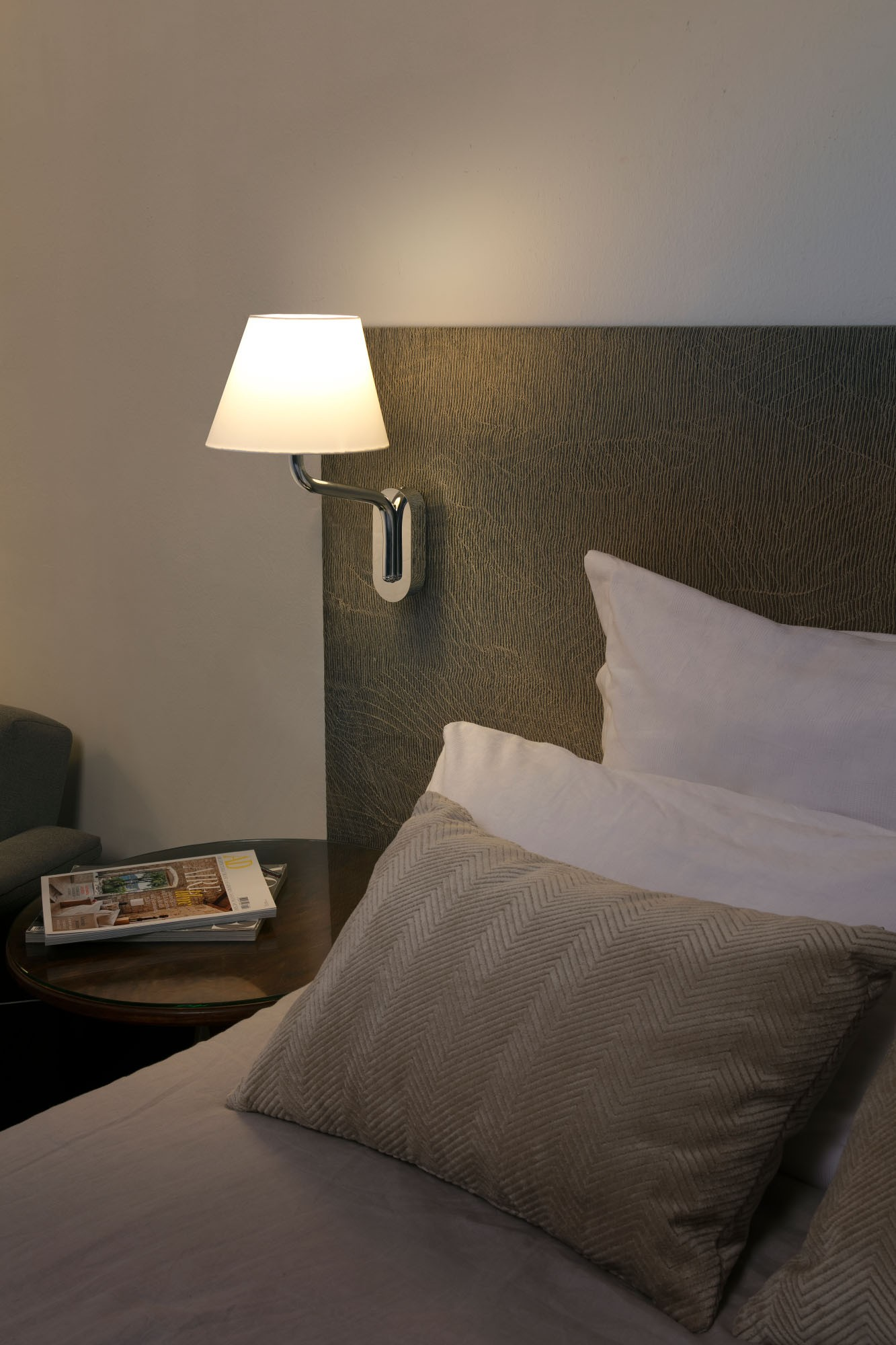 Un comodino con riviste accanto a un letto con cuscini e una lampada da parete.