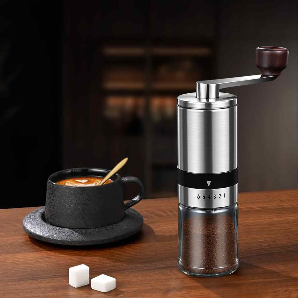 Kaffeemühle aus Metall mit Holzgriff neben einer Tasse Kaffee und zwei Zuckerwürfeln.