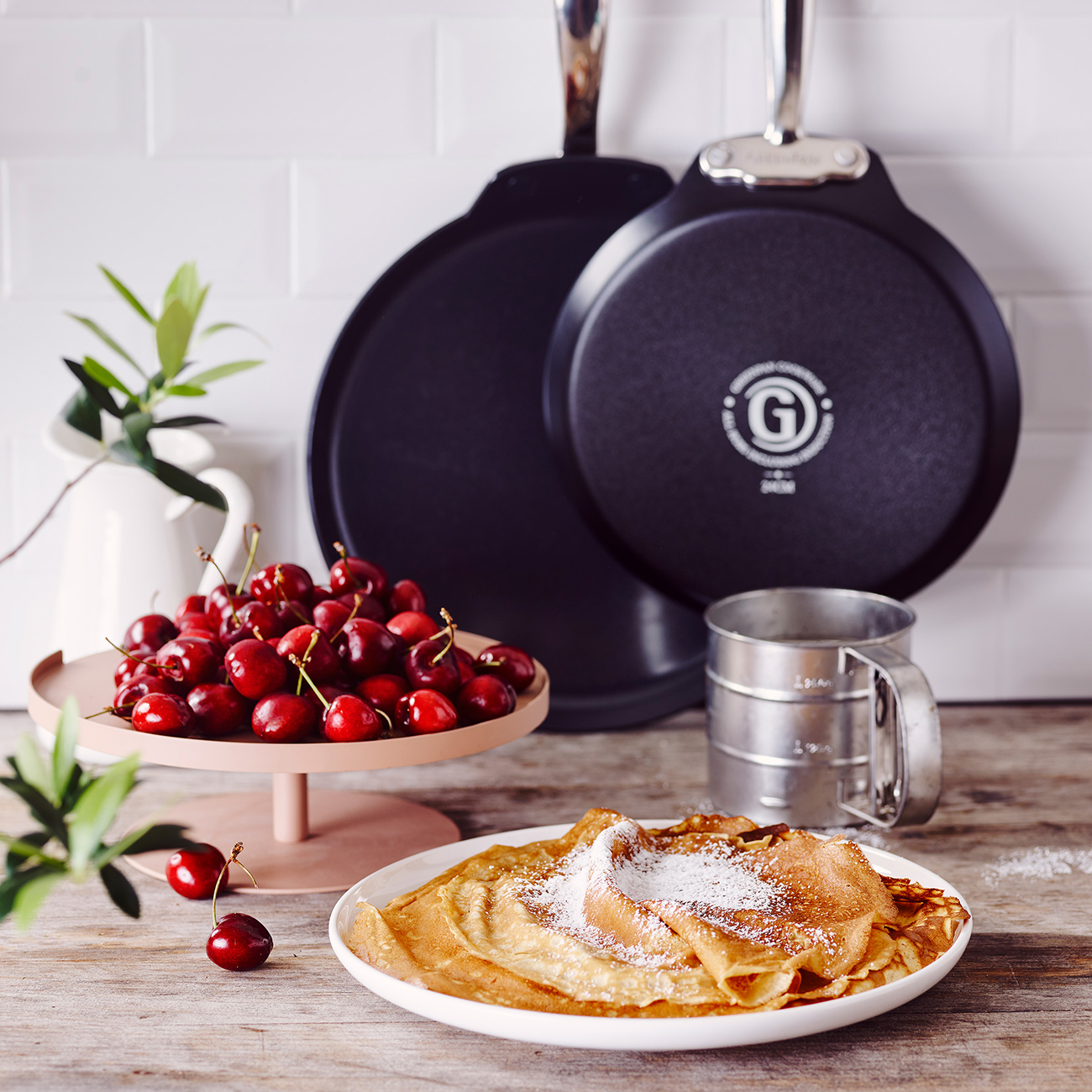 Ein Teller Crêpes mit Puderzucker bestäubt, neben Kirschen und Pfannen.
