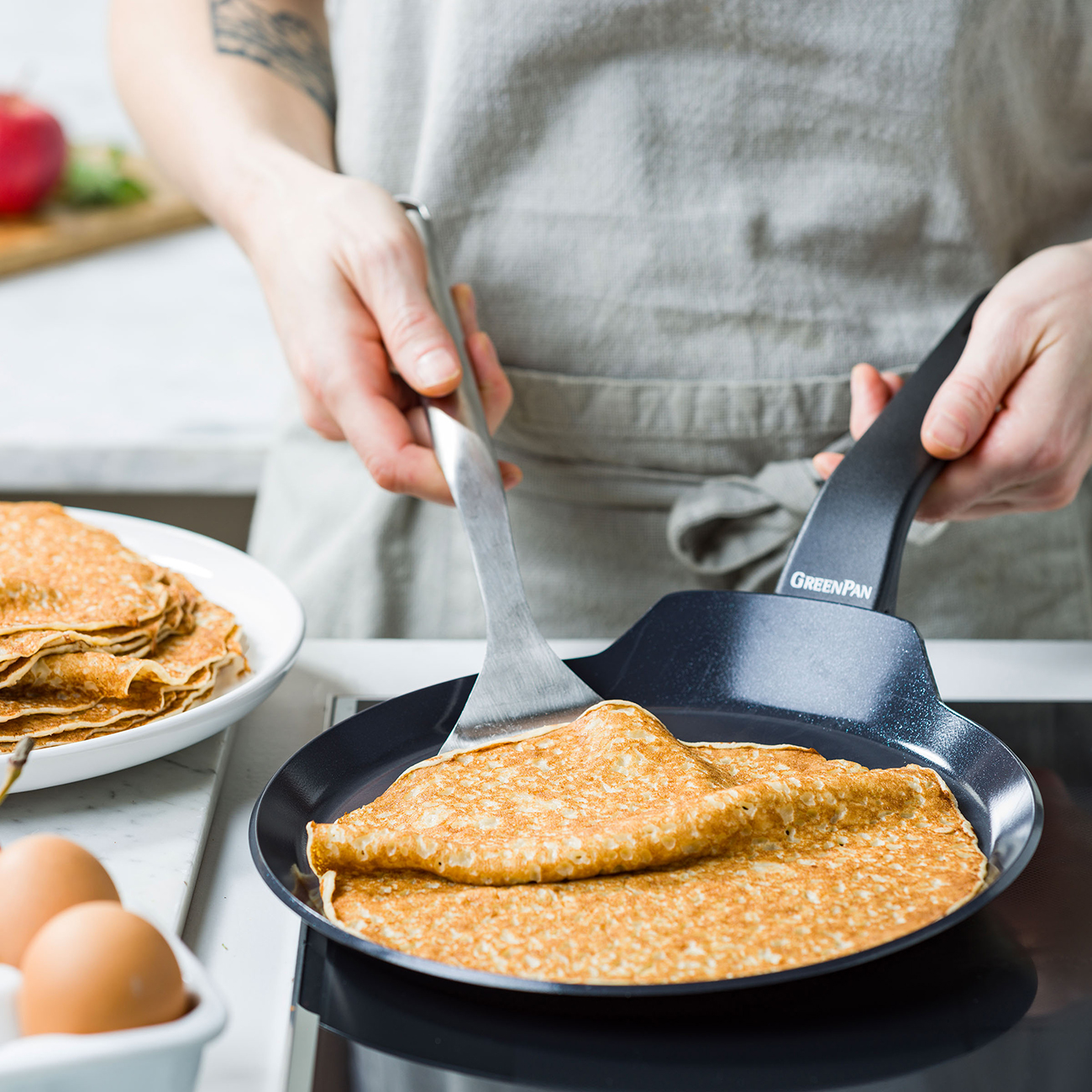 Eine Person in einer Schürze wendet einen Crêpe in einer GreenPan-Pfanne. Gestapelte Crêpes und Eier in der Nähe.