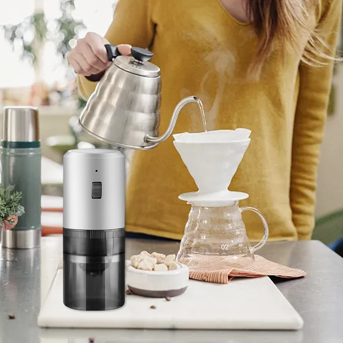 Eine Person gießt heißes Wasser aus einem Wasserkocher in einen Kaffeefilter. Eine Kaffeemühle steht daneben.