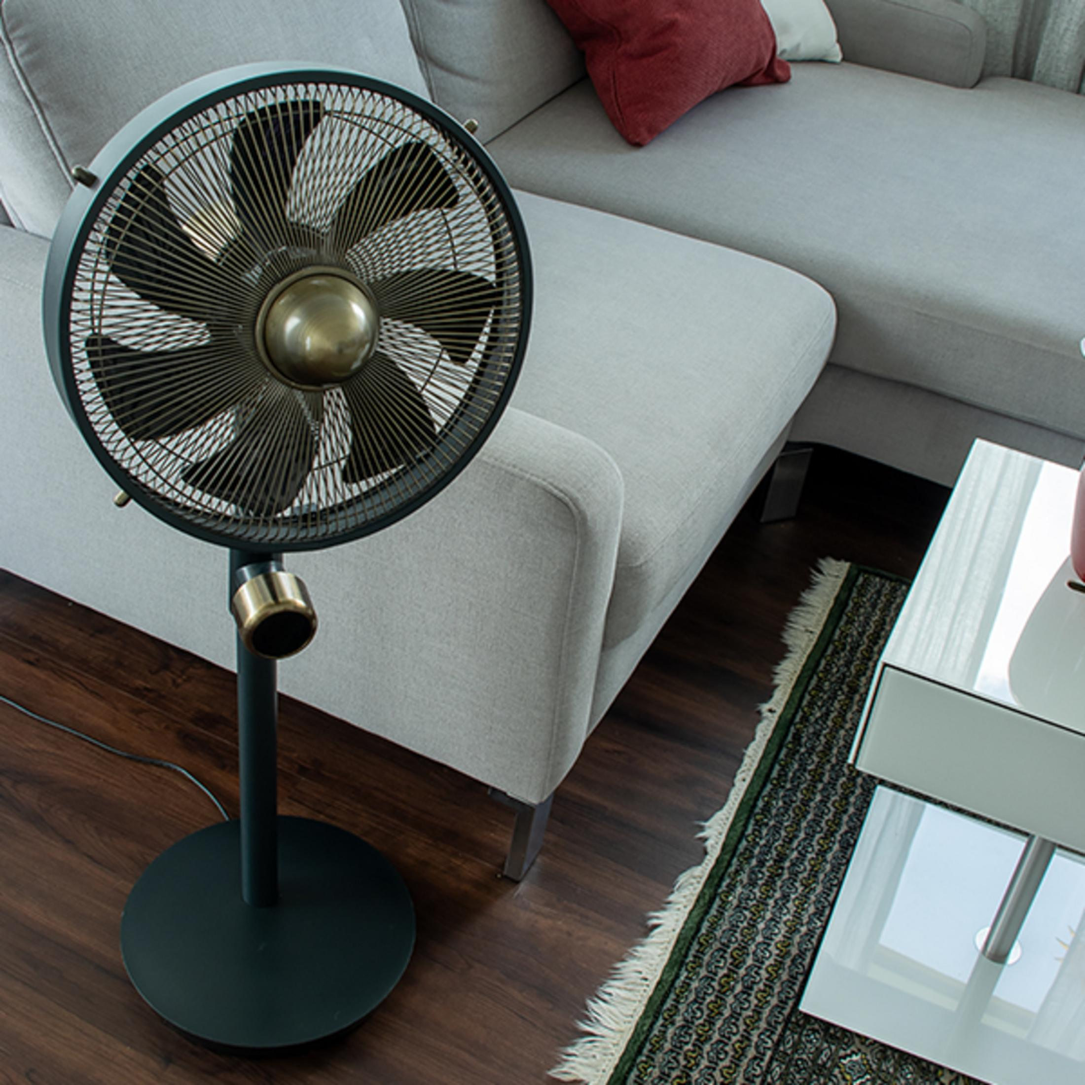 Un ventilateur sur pied noir est placé à côté d'un canapé gris avec coussins.