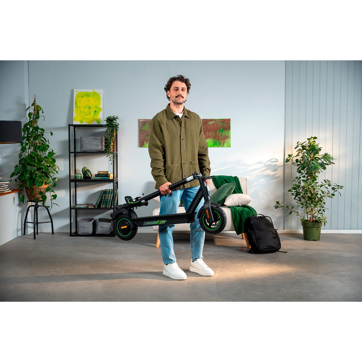 Hombre sujetando un patinete eléctrico plegado en sala con plantas y muebles.